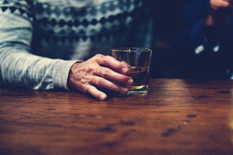 A man holds a tumbler of a dark alcohol, set against a wooden bar.